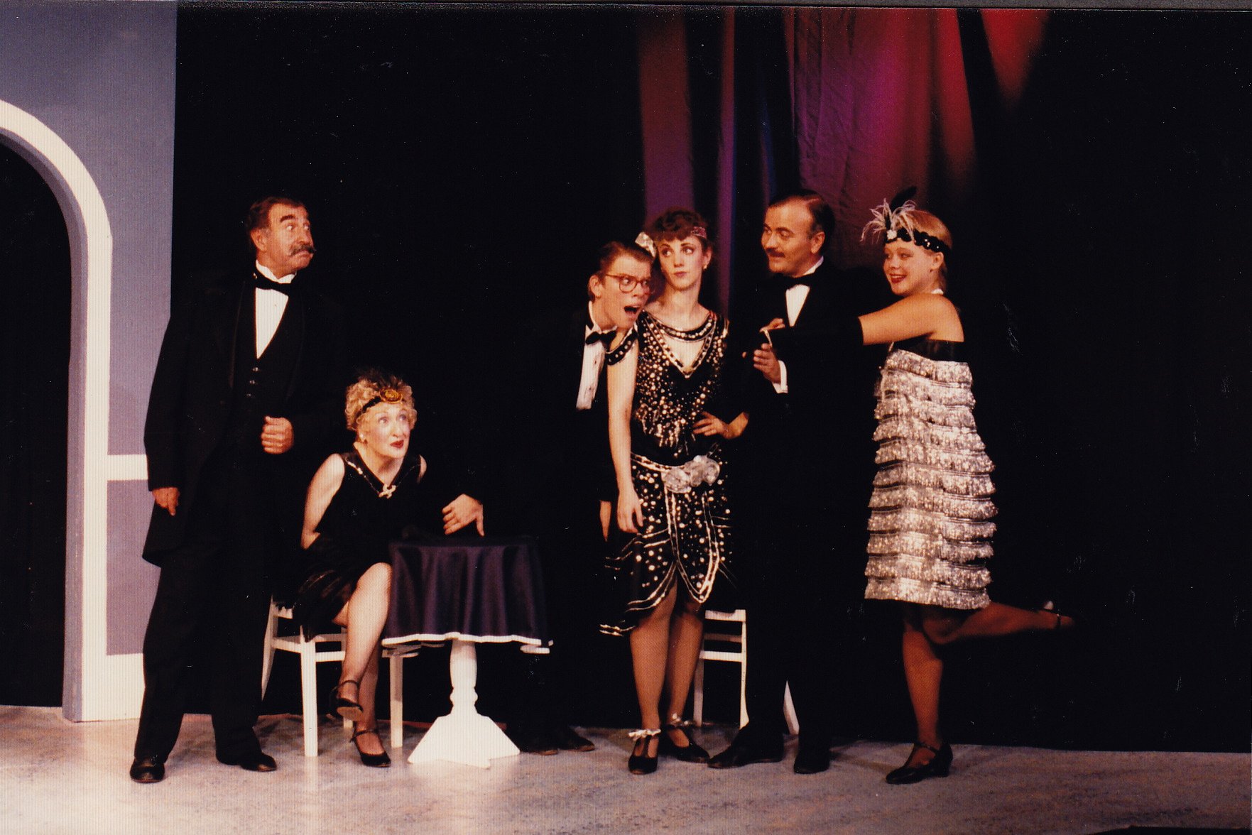 Group of people dressed in 1920s-style costumes with flapper dresses and tuxedos, standing on a stage with a dark background.