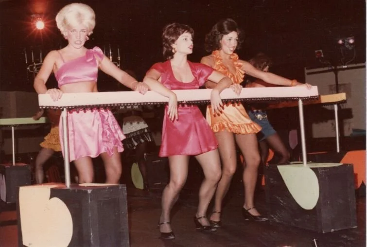 Three women in 1960s-style dance costumes performing on stage, with colorful outfits and a retro setting.