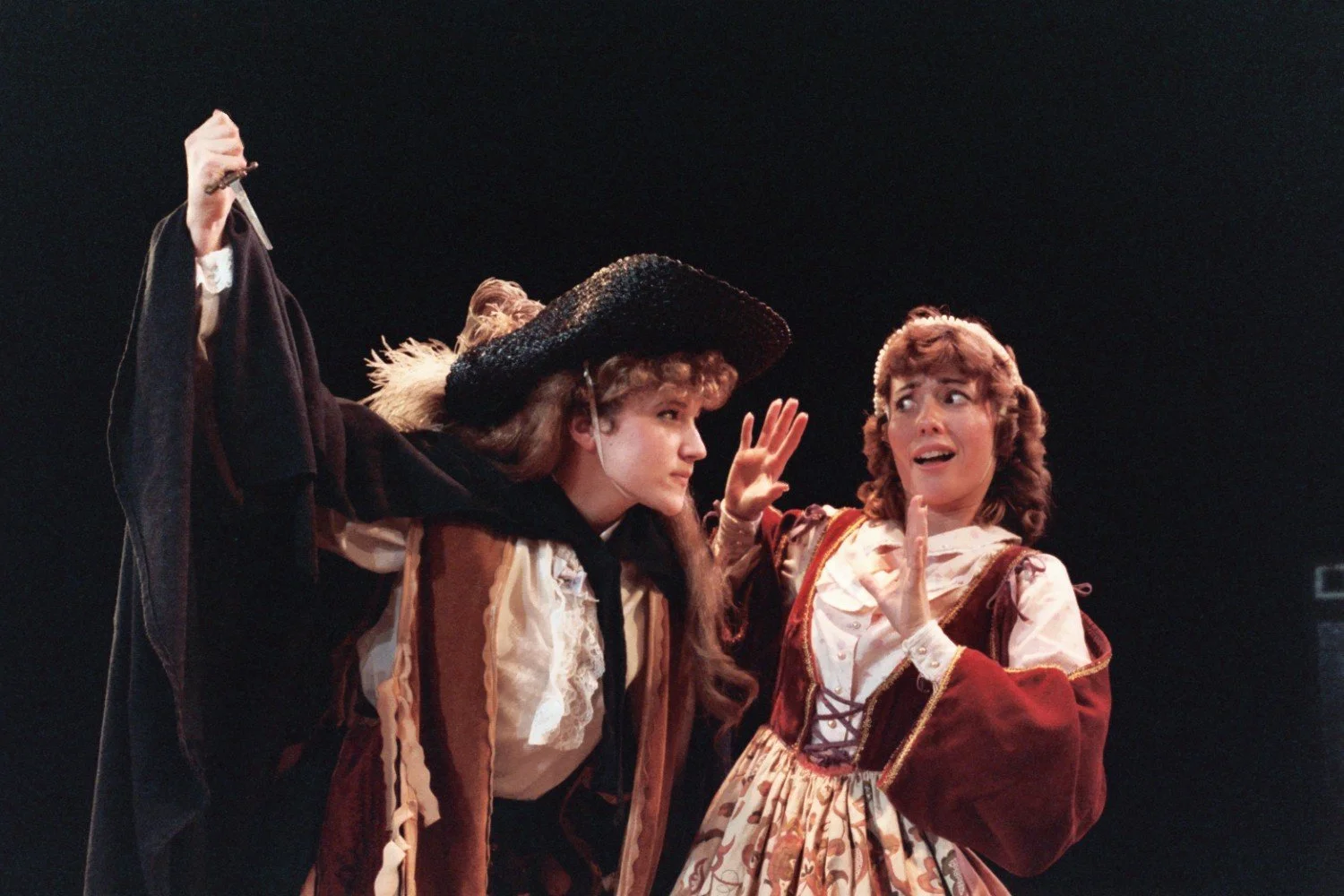 Two actors in theatrical costumes, one holding a dagger, performing a dramatic scene on stage.