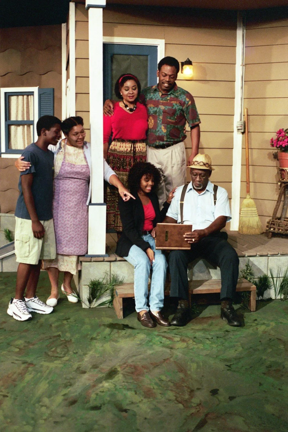 Family gathered on a porch, with an older man sitting and holding a book. Other members are standing and sitting around him.