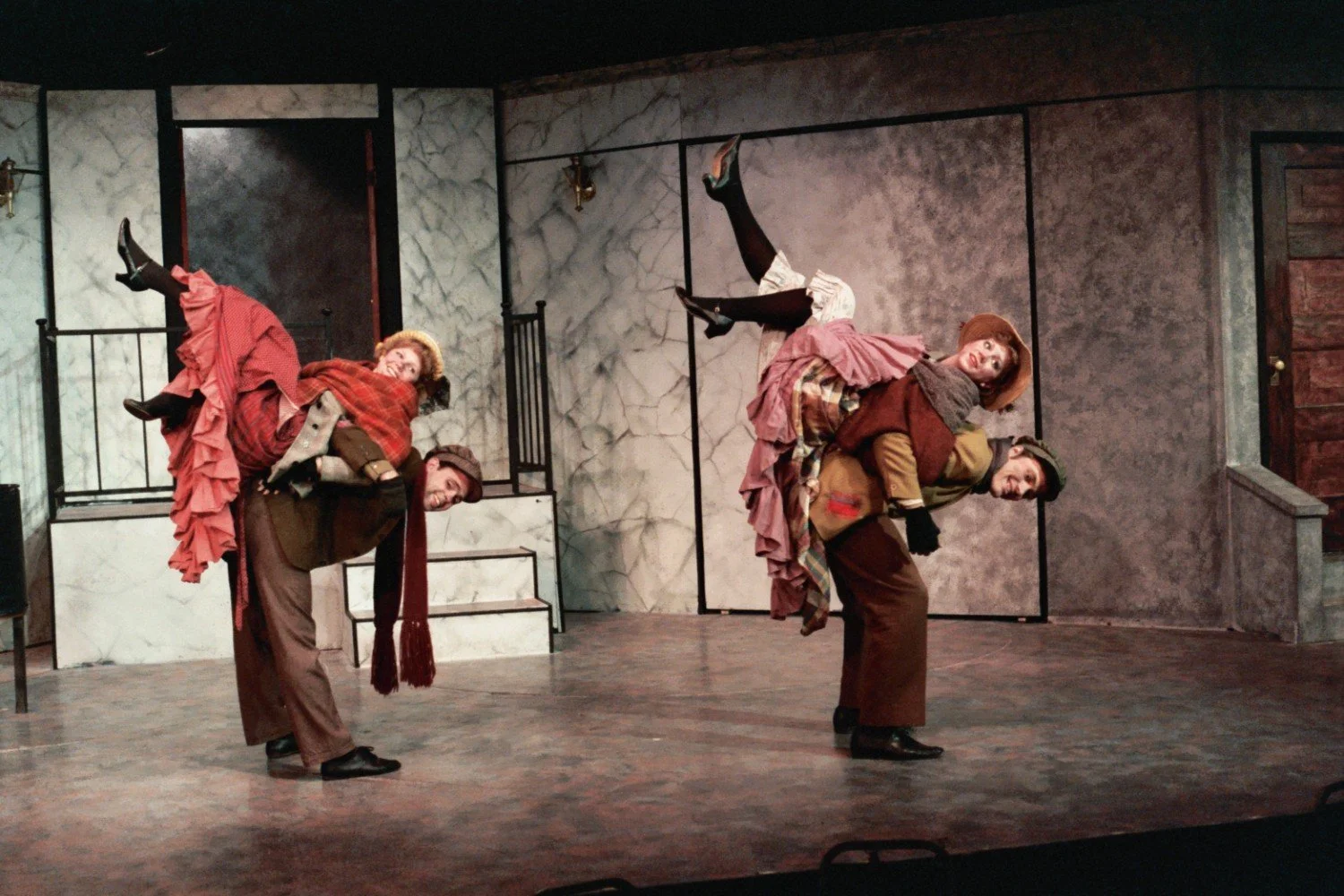 A theatrical performance with two male actors in costume carrying two female actors on their backs, set against a stage with a Victorian-style backdrop.