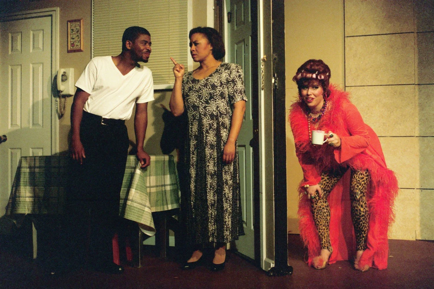 Three people in a theatrical scene; one person in a white shirt leans on a table, another in a patterned dress gestures seriously, and a third in a vibrant red costume with leopard print accents and curlers holds a cup.