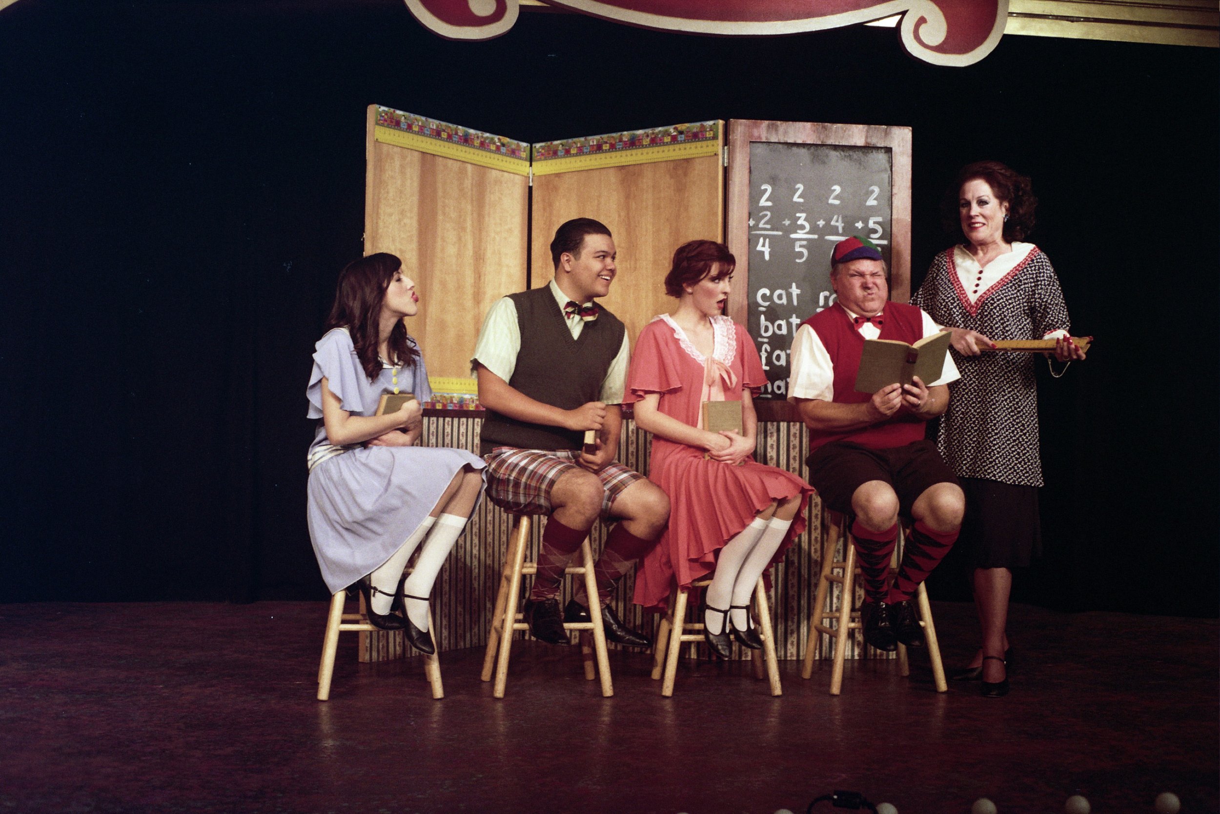 A theatrical performance scene with four actors dressed in vintage school attire sitting on stools, and a woman dressed as a teacher holding a pointer. A chalkboard with simple math problems and words is in the background.