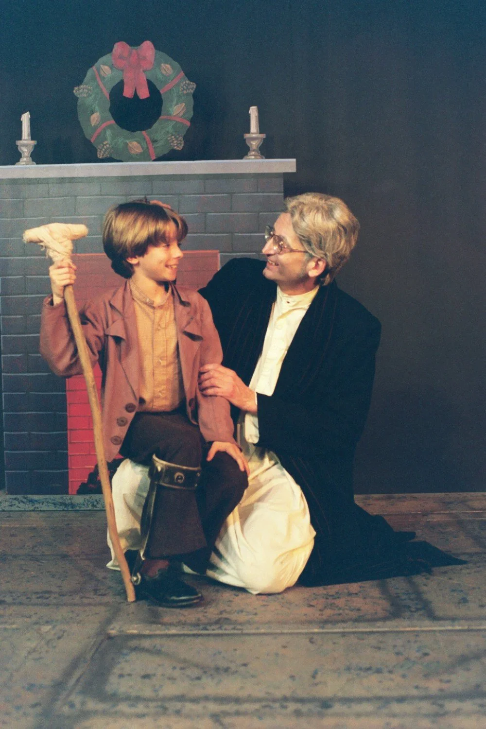 A theater scene with two people, likely from "A Christmas Carol," depicts an older man in glasses and a child with a crutch, set against a fireplace with a wreath.