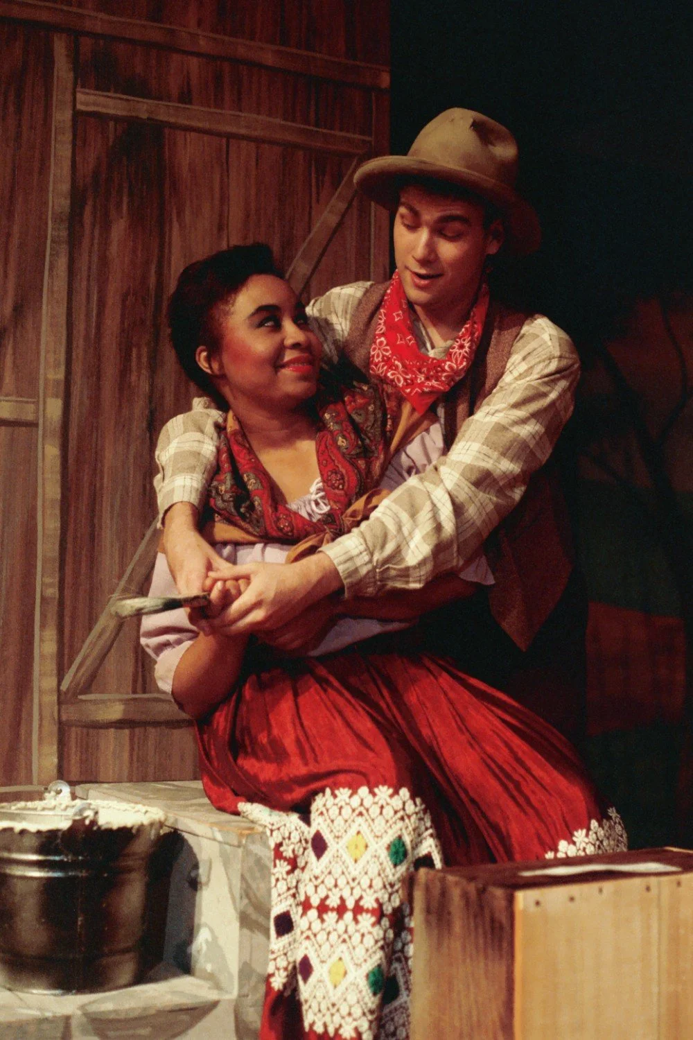 Two people in a theatrical scene, dressed in period Western costumes. The man is wearing a cowboy hat, red bandana, and plaid shirt, embracing a woman in a red skirt and patterned apron. They appear to be engaged in a warm, affectionate moment on sta