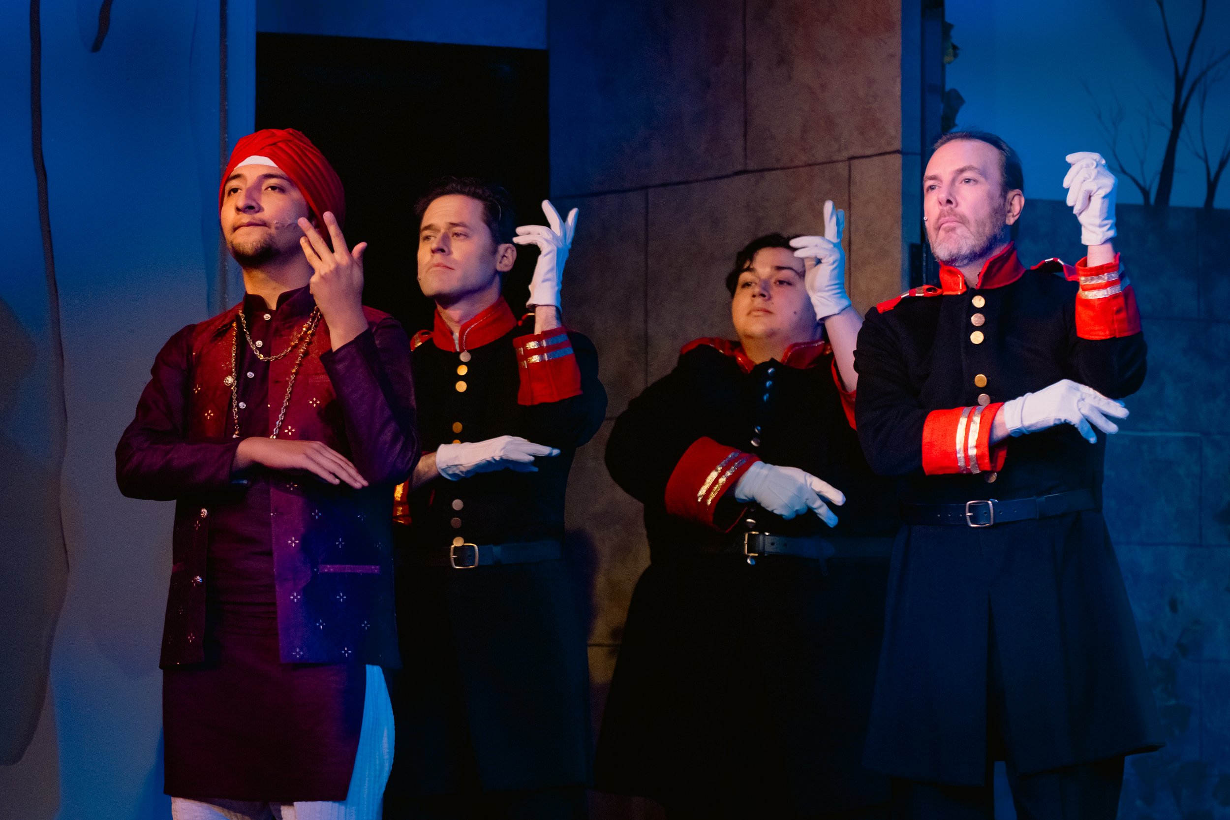 Group of four actors on stage, two wearing military-style costumes with red accents, black gloves, and the other two in cultural attire with a red turban. They are making expressive hand gestures and have serious expressions.