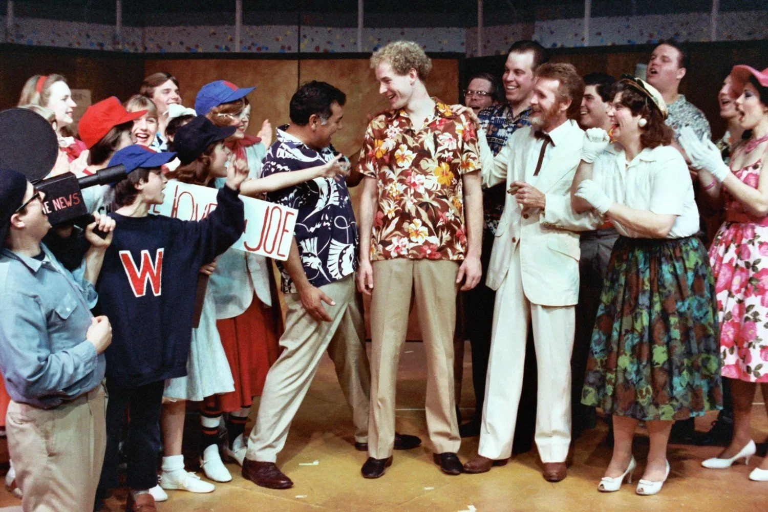 Group of people wearing colorful, vintage-style clothing; some holding signs that say 'W' and 'We Love You Joe'; includes a person with a news camera prop.