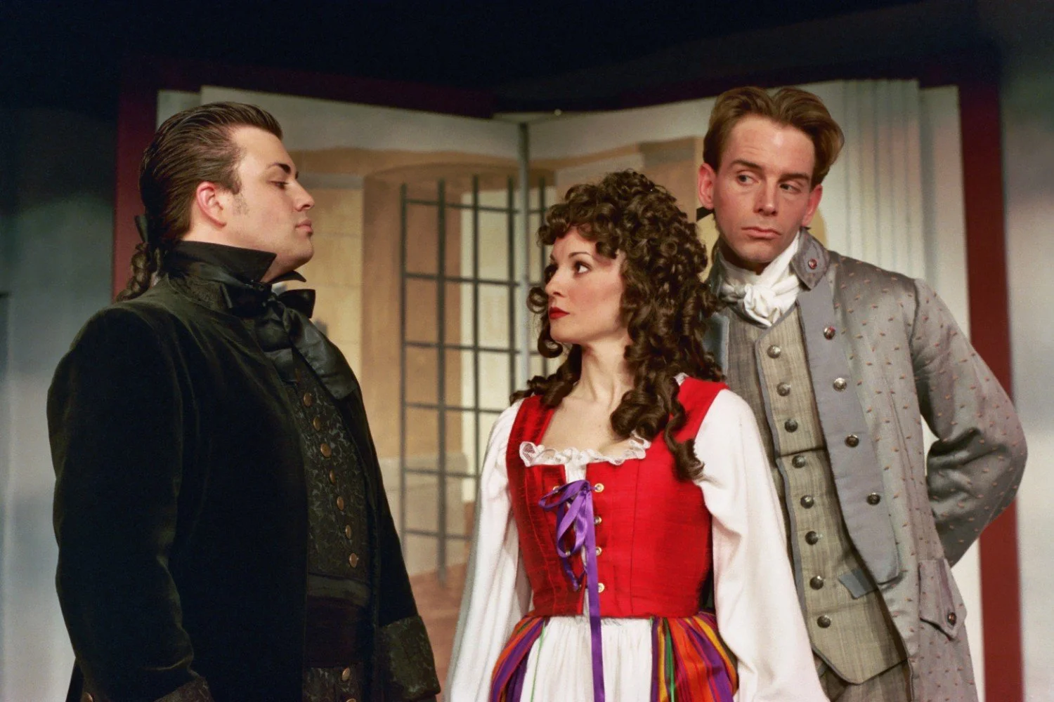 Three actors in period costumes on stage during a theatrical performance, wearing ornate and historically styled clothing. The scene depicts a dramatic interaction among them.