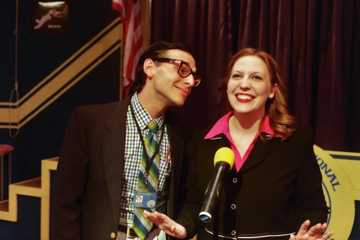 Two people wearing business attire, standing in front of a microphone on a stage. The man is wearing glasses and a checkered shirt with a tie, while the woman is smiling and wearing a blazer. An American flag and stage decorations are in the backgrou