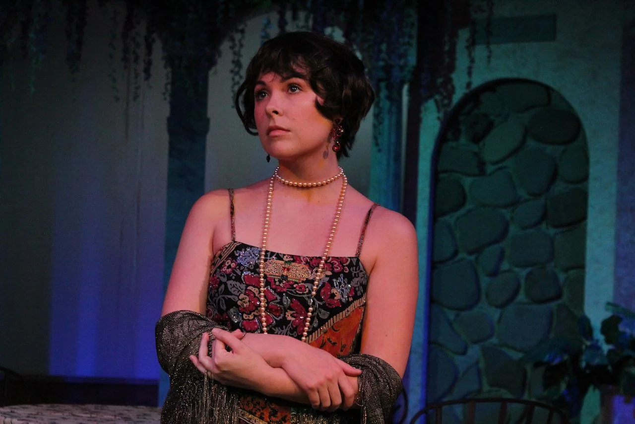 Woman in vintage floral dress with pearl necklace, standing in a dimly lit room with stone wall background.