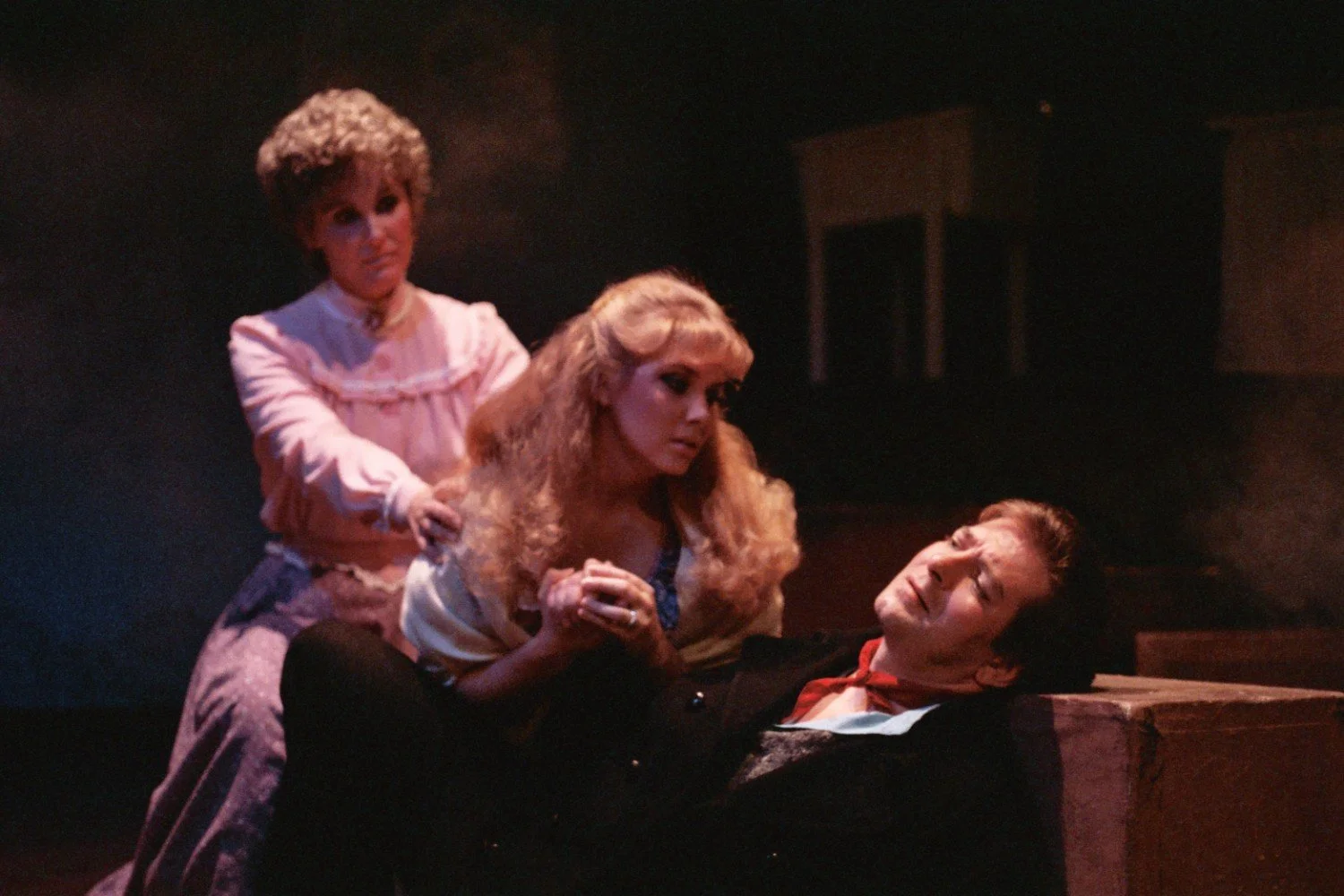 Three actors in period costumes performing a dramatic scene on stage, with one lying down appearing wounded and two others showing concern.