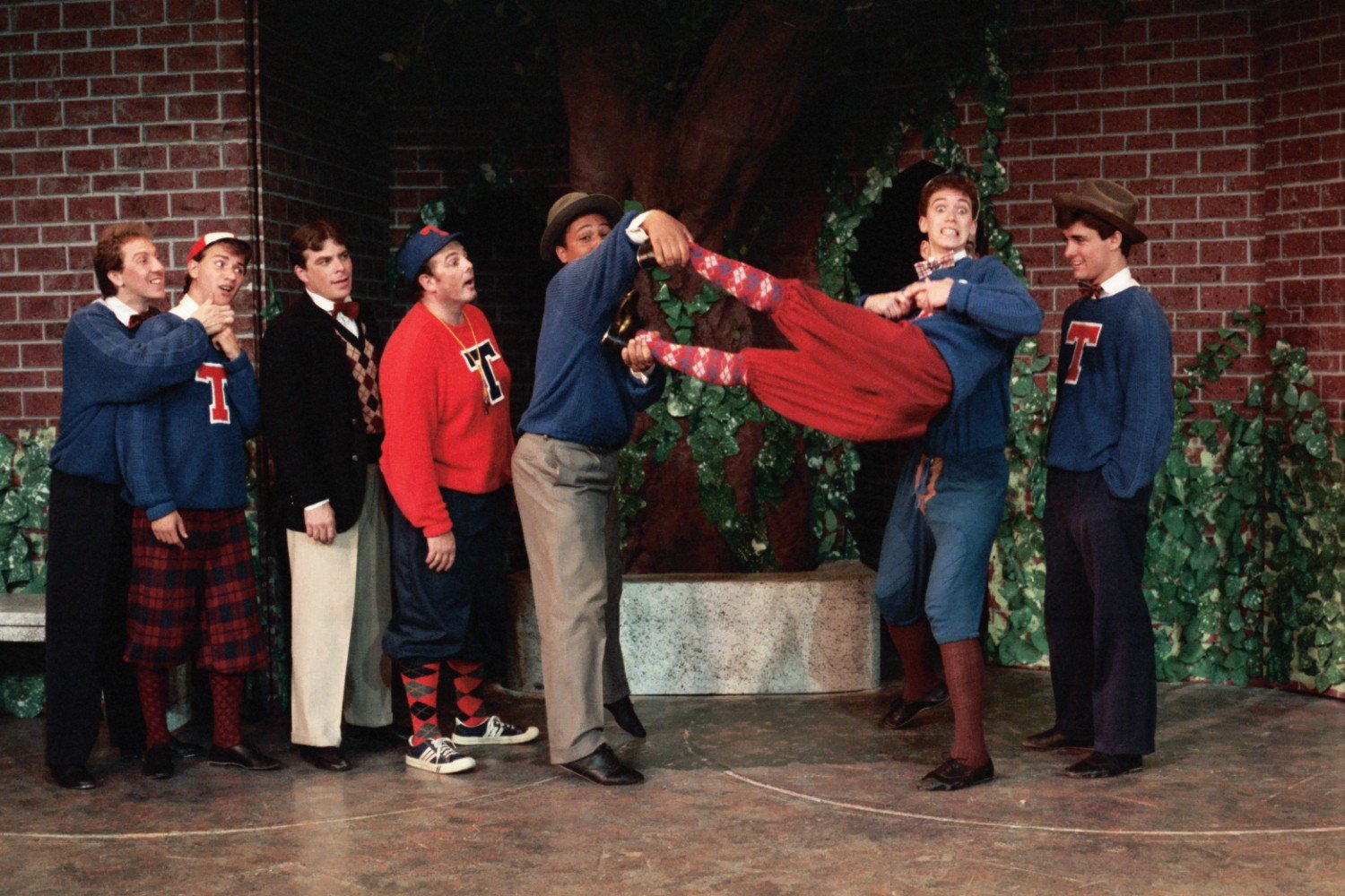 Group of people in theatrical costumes, with some wearing blue and red sweaters with the letter 'T', engaged in a humorous scene, two individuals holding another person horizontally. Background includes a brick wall and greenery.