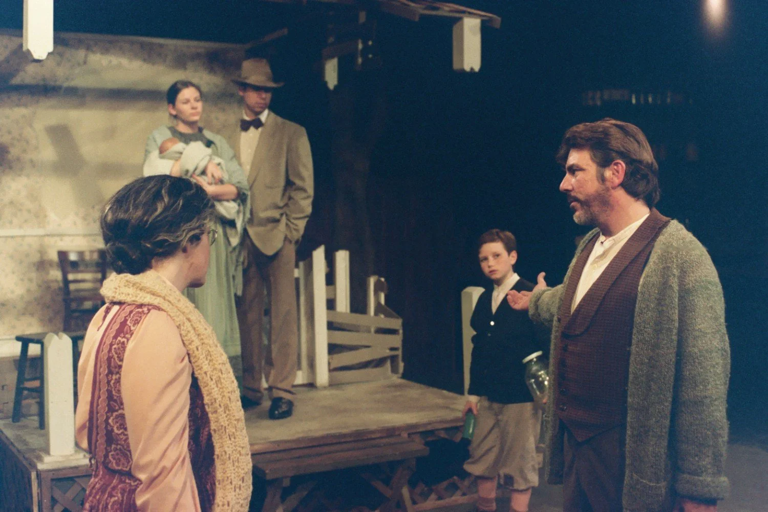 Stage scene with five actors in period costumes, including a man in a sweater, a woman in a scarf, a couple holding a baby, and a boy, in a rustic setting with a wooden porch backdrop.