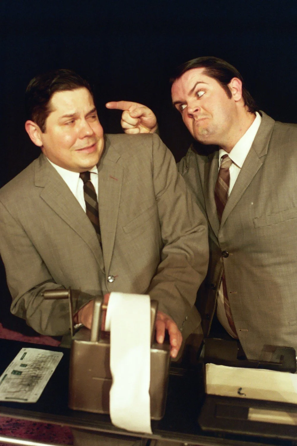 Two men in suits, one with an expressive gesture, and a vintage-style adding machine with paper roll.