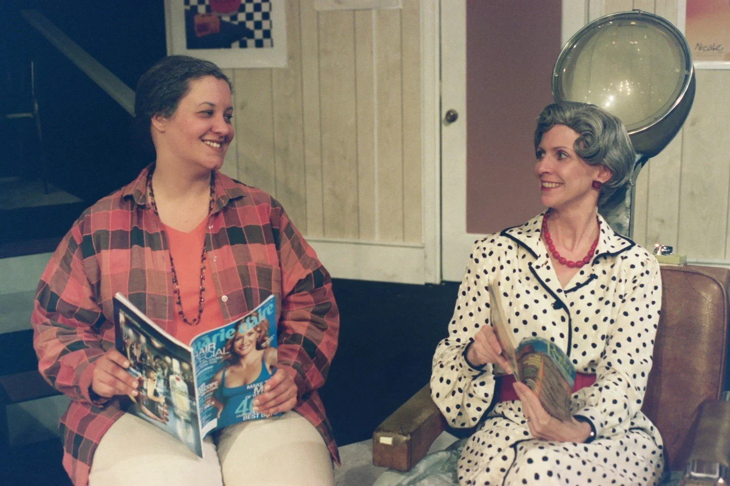 Two women sitting in a salon, one in a polka dot dress under a hair dryer, holding a magazine, and the other in a plaid shirt, holding "Marie Claire". They are smiling at each other.