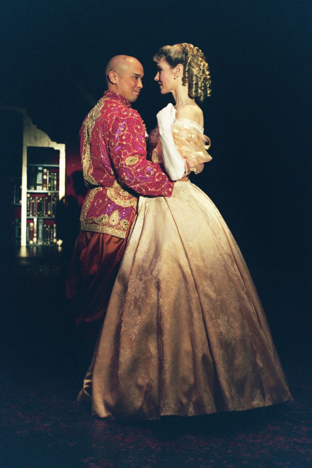 A man and woman in elegant, period costume are dancing together on stage. The man wears an ornate red and gold outfit, while the woman wears a luxurious gold gown. They are facing each other, creating an intimate moment.