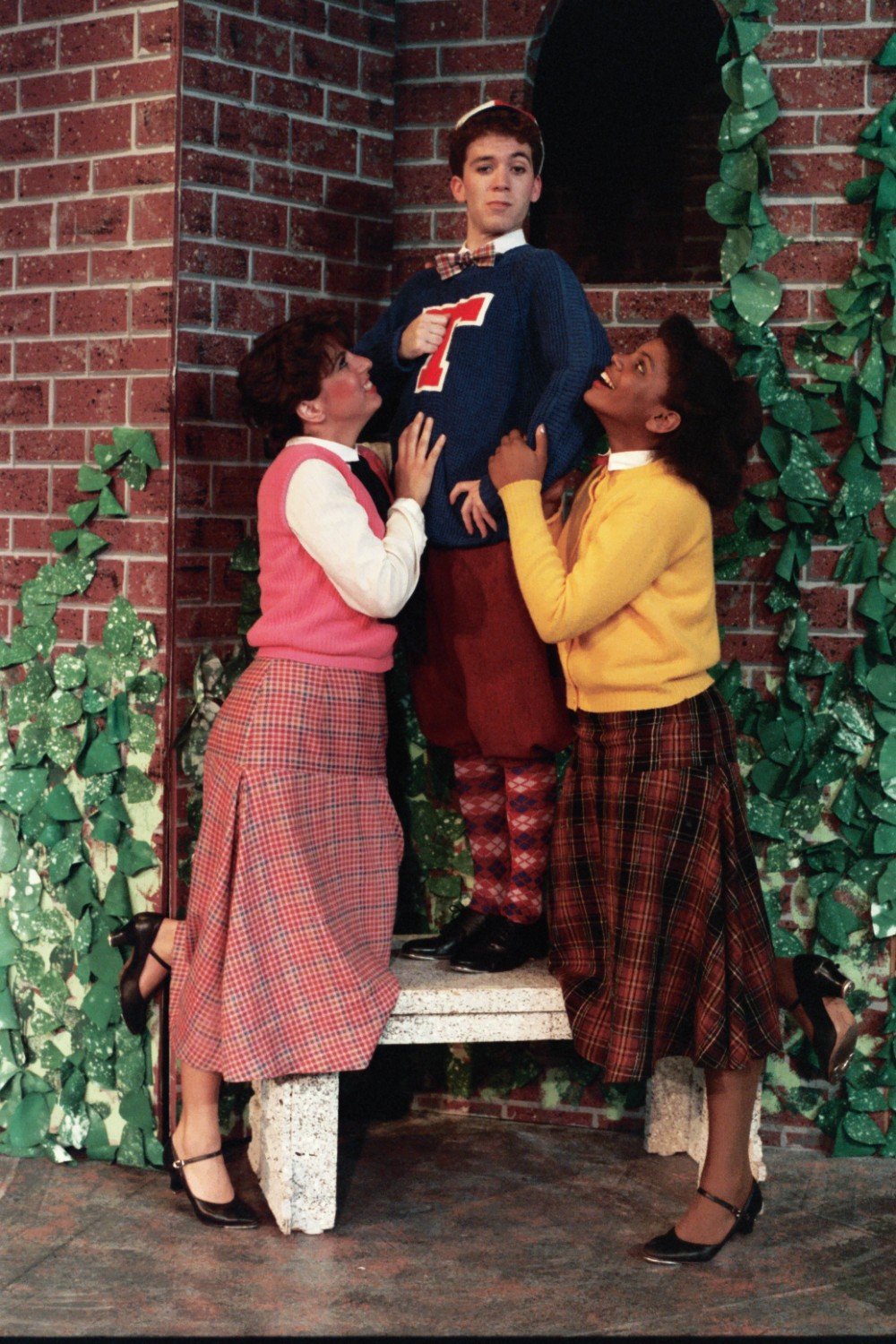 Three people in vintage-style clothing on a theater set with brick background and greenery. Two women in skirts and sweaters are admiring a person in a letterman sweater and cap, standing on a bench.