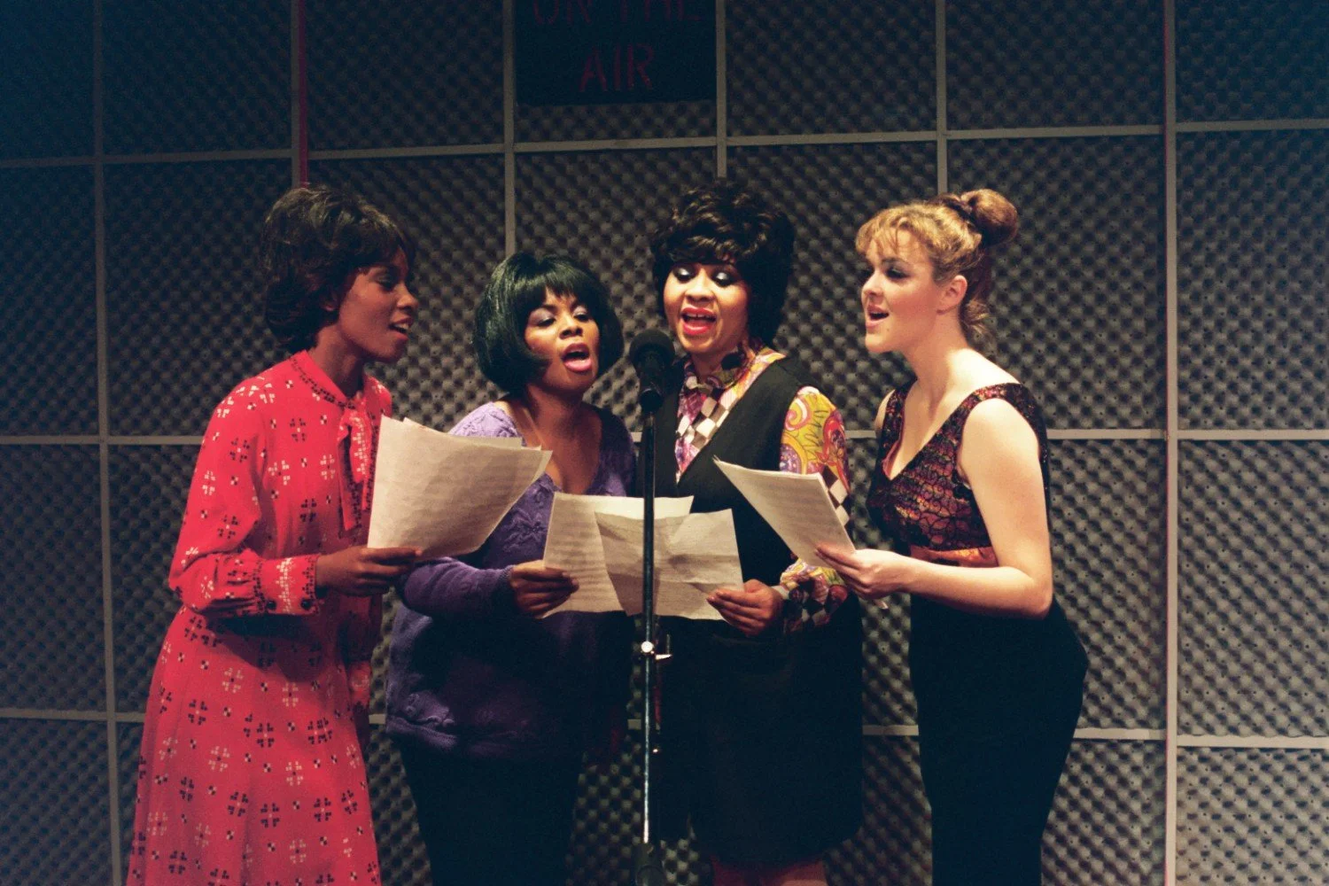 Four women singing into a microphone in a recording studio, holding papers with soundproofing panels in the background.