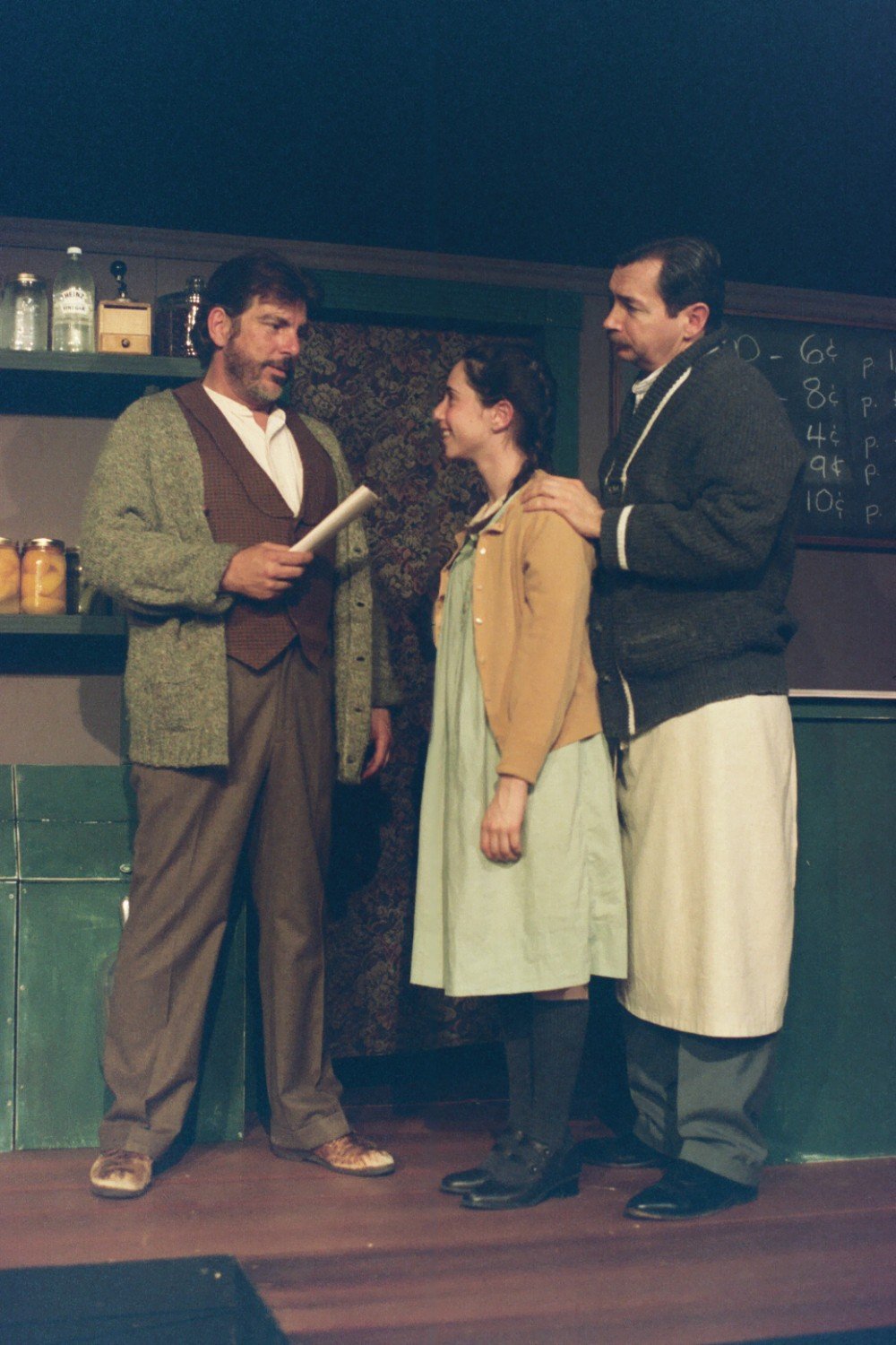 Three actors performing on stage, portraying a family scene in a play. The man on the left holds a document, the woman in the middle looks at him smiling, and the man on the right has his hand on her shoulder. The set includes shelves with jars and a