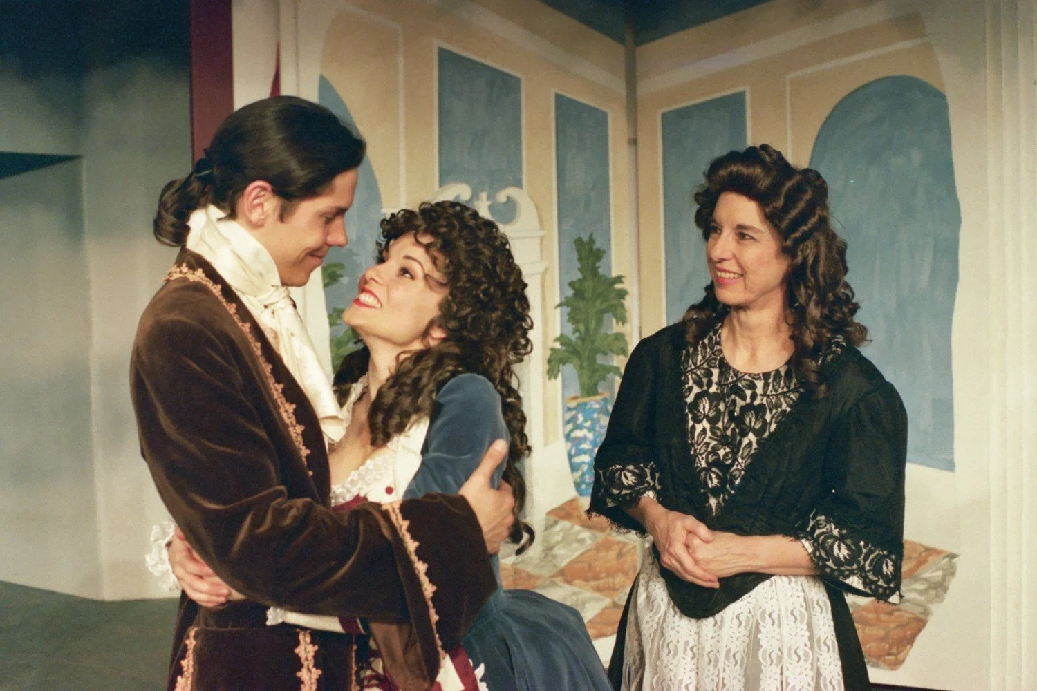 Three people in historical costumes on stage, two embracing and one smiling, with a painted backdrop.