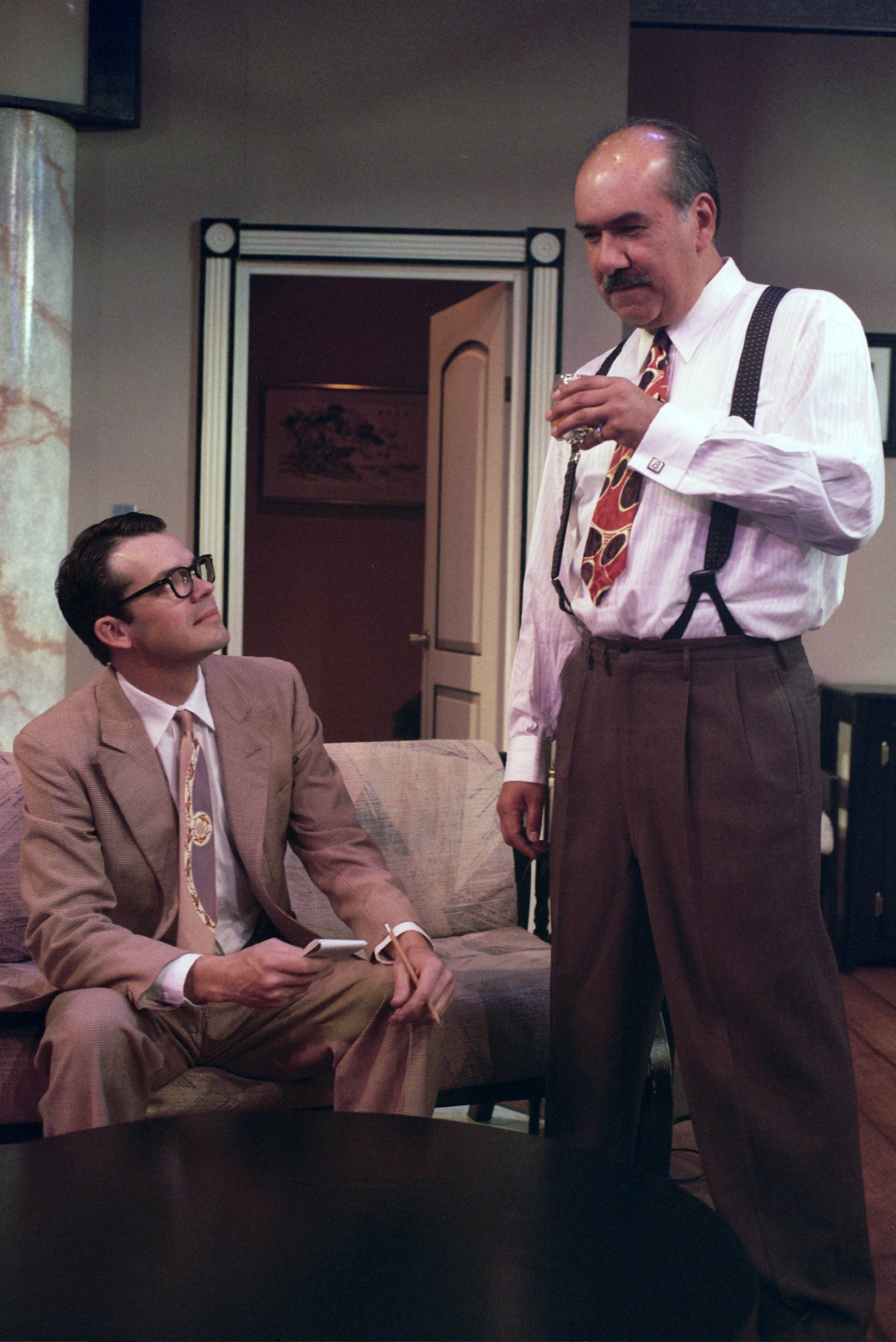 Two men in a living room, one seated holding a paper, the other standing with a drink.