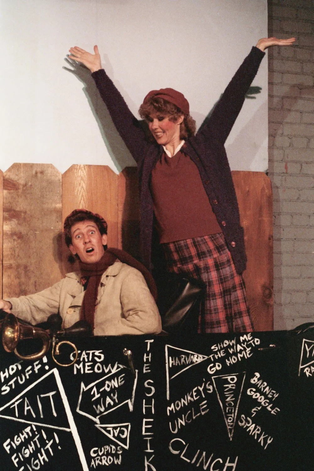 Two people in period costumes, a man seated with a trumpet and looking surprised, and a woman standing with arms raised, next to a fence with painted signs and pennants on a table.
