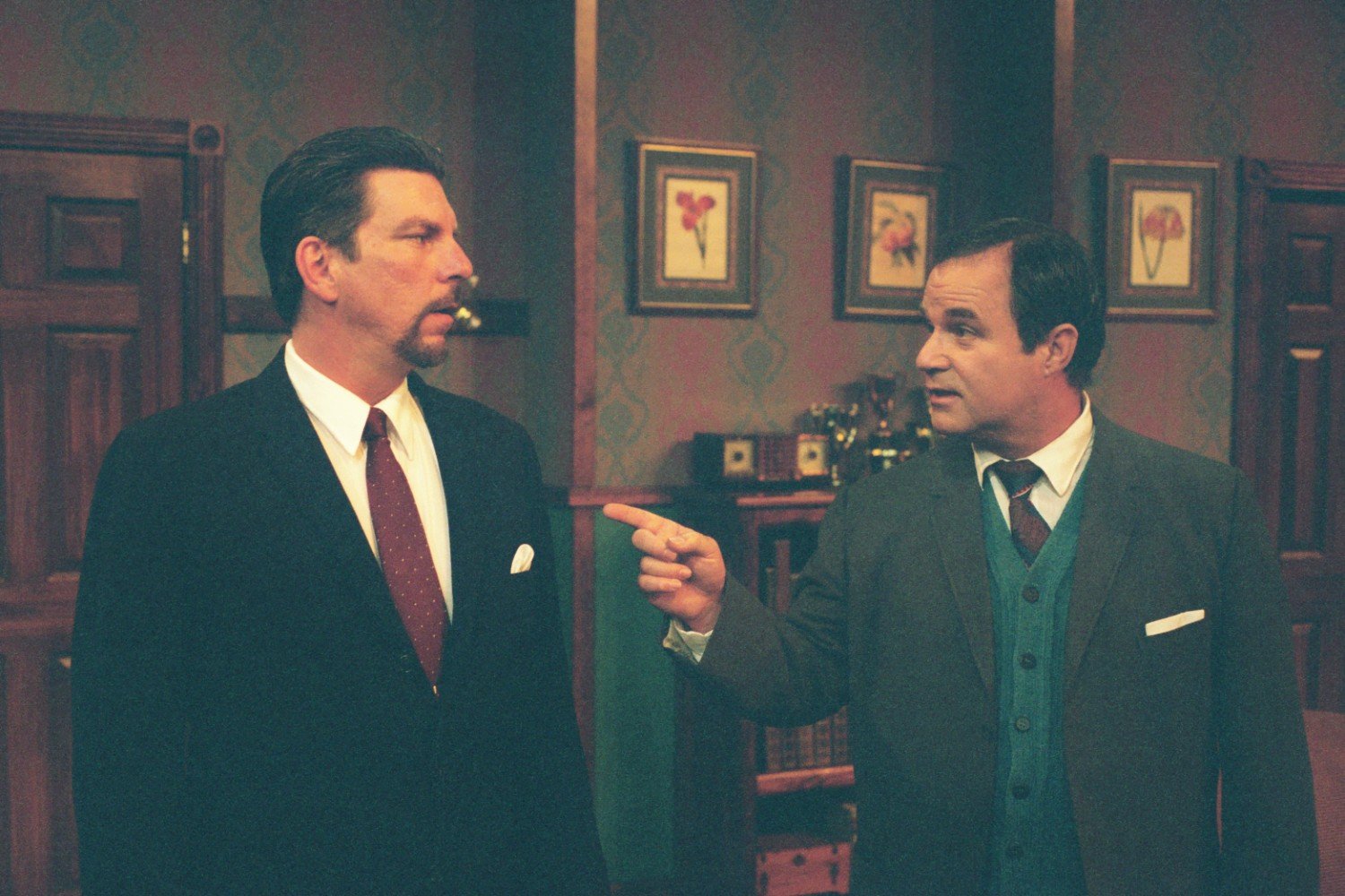 Two men in suits having a conversation in a room with framed floral paintings and wooden doors.
