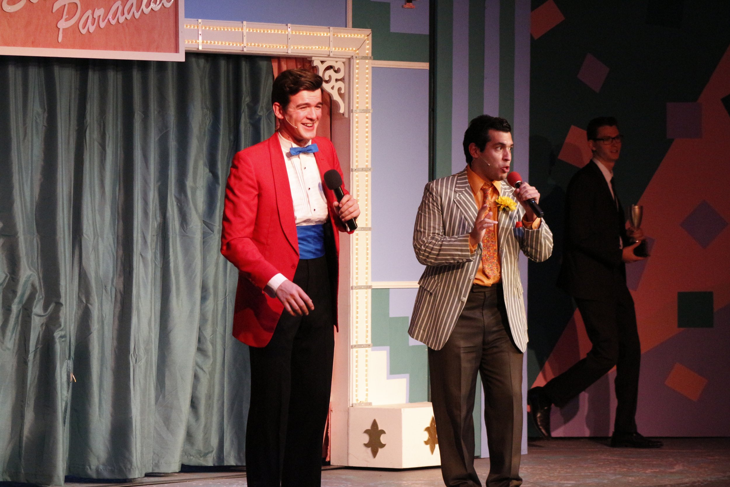 Two men performing on stage, one in a red jacket and the other in a striped suit, each holding a microphone, with a stage setup in the background including a curtain and decorative frame.