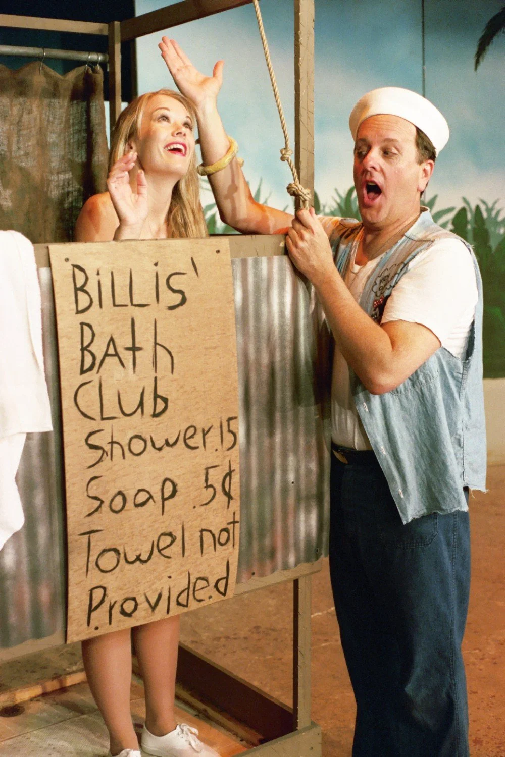 A theatrical scene with a woman in a makeshift shower stall and a man in a sailor costume. The stall has a sign reading "Billis' Bath Club Shower 15¢ Soap 5¢ Towel not Provided." The backdrop resembles a tropical setting.
