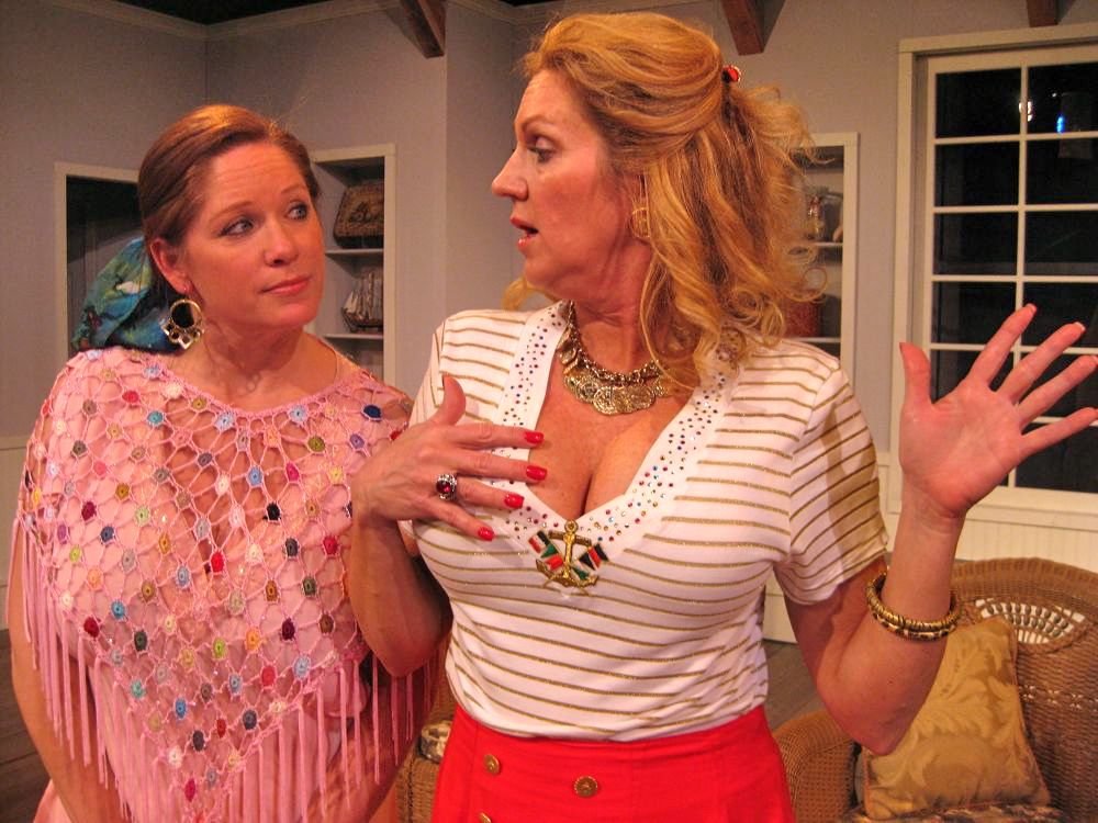 Two women having a conversation indoors, one wearing a pink crochet top with colorful accents, and the other in a white and gold striped shirt with a red skirt. They appear to be in a living room with beige furniture.