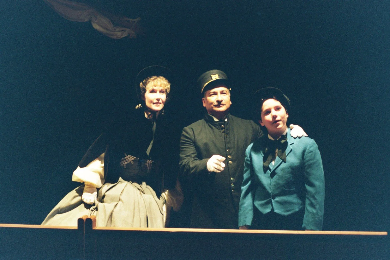 Three people dressed in historical clothing, possibly Victorian-era, standing close together and looking forward. One person is wearing a military-style uniform and cap, while the others are in formal dress with hats.