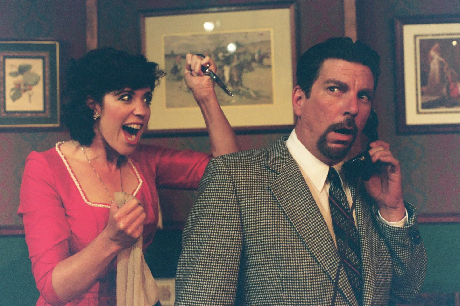 A woman in a red dress holding scissors behind a man on the phone in a vintage room.