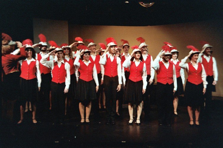Group of performers on stage in red vests and hats with red feathers.