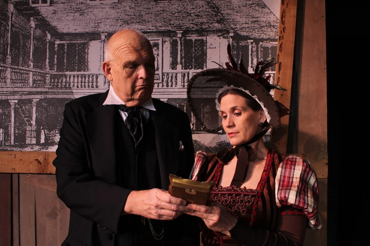 Two people in 19th-century attire examining a small book or document, set in front of a historical building backdrop.