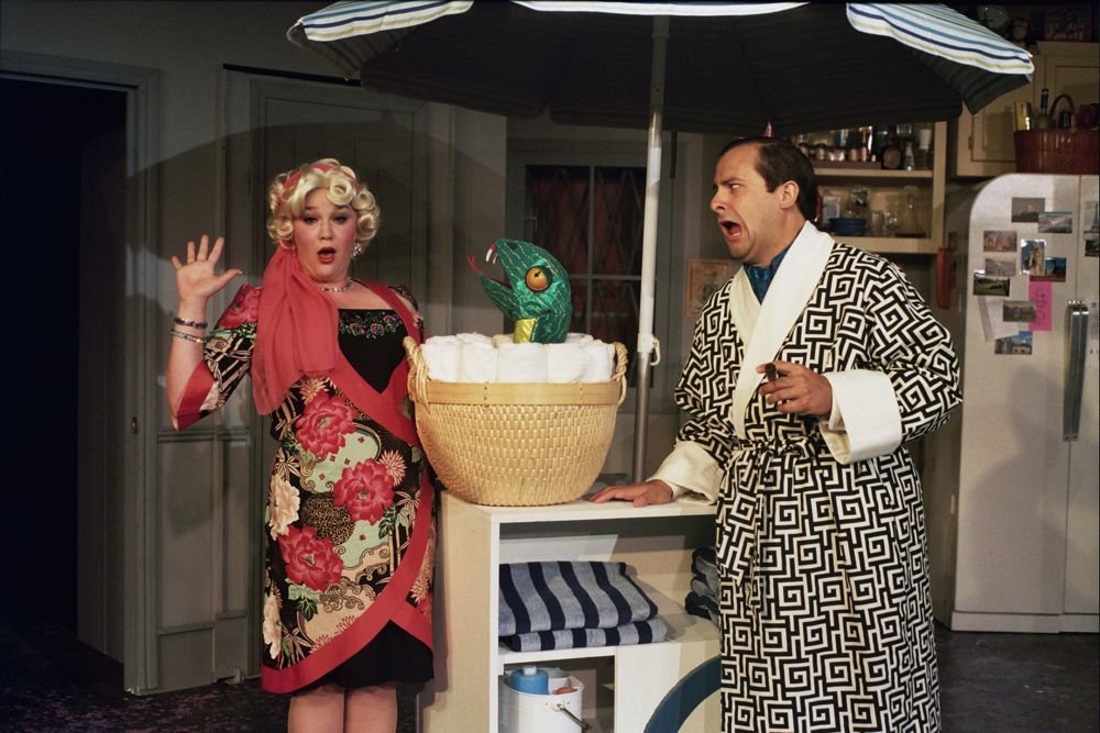 Two people in a kitchen scene, both looking surprised. A person in a floral dress and a man in a geometric-patterned robe react to a green puppet emerging from a basket on a table, under an umbrella.