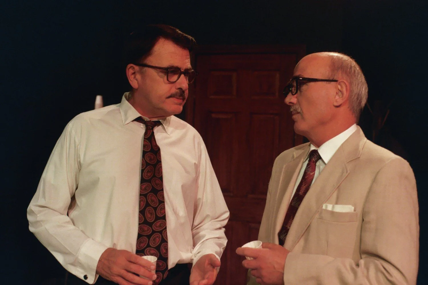 Two men in suits having a conversation with each holding a cup indoors.