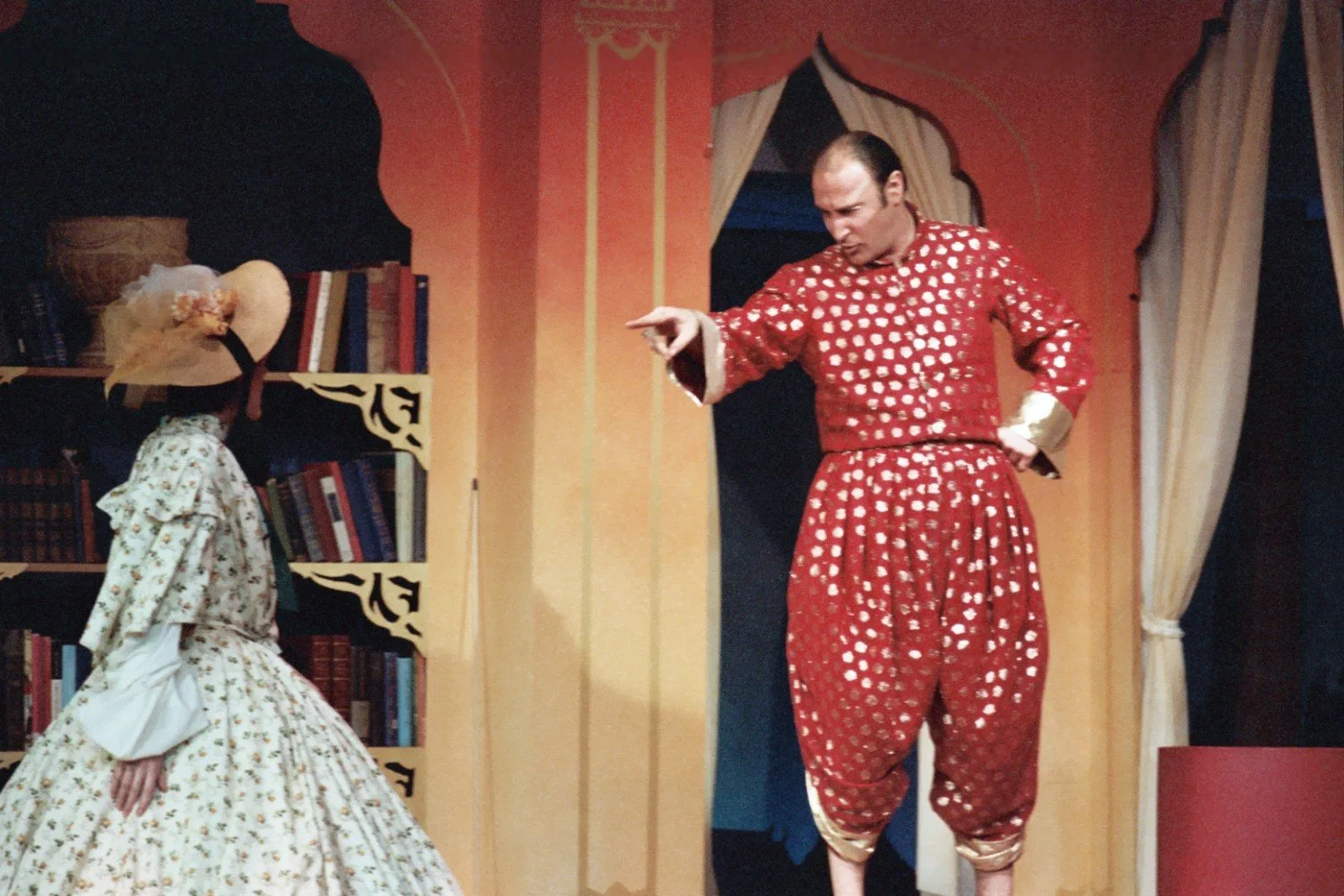 A scene from a theatrical performance featuring a man in a red costume with gold patterns pointing towards a woman in a floral patterned dress and large hat, standing in front of a bookshelf.