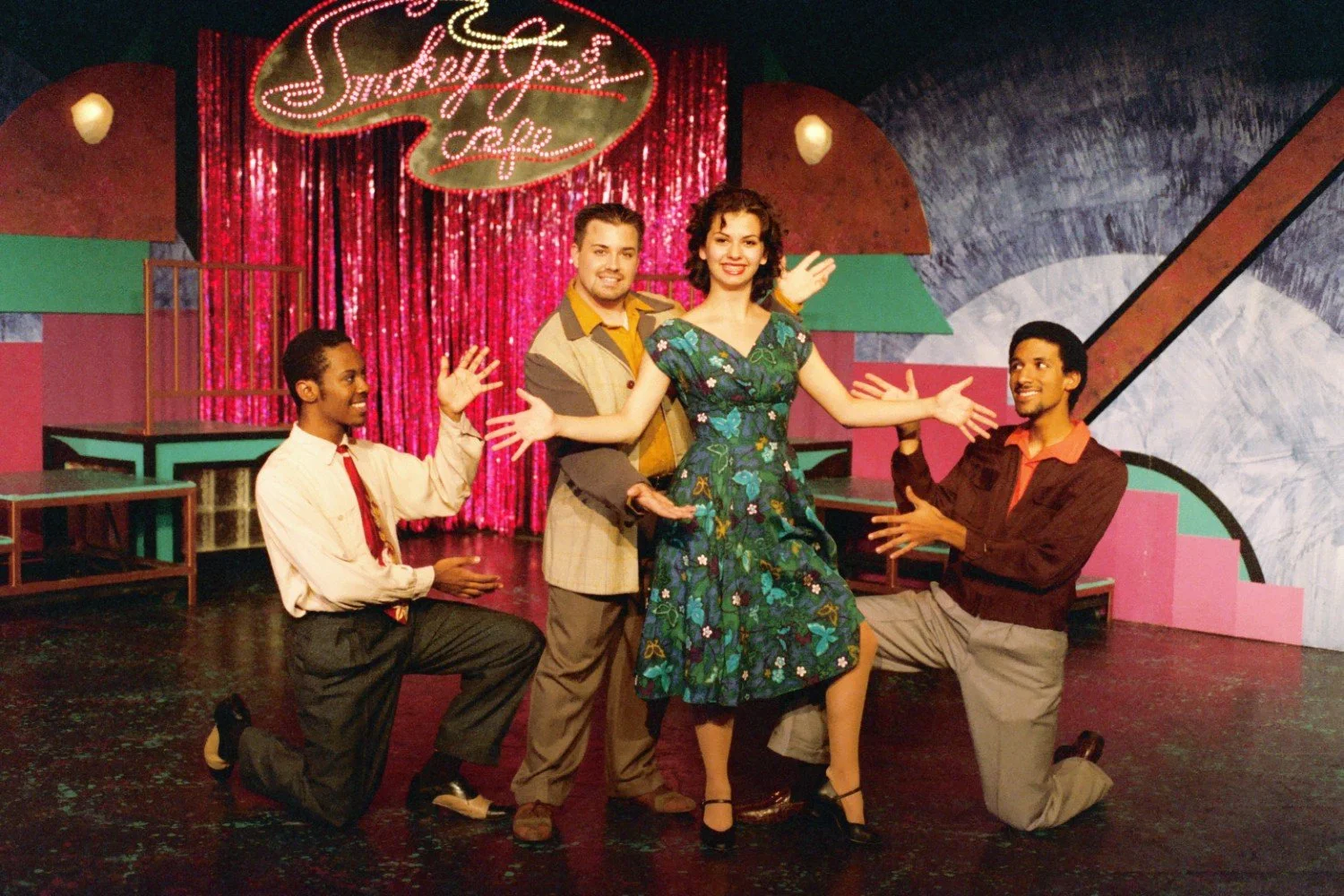 Four performers on stage with a "Smokey Joe's Cafe" sign in the background, wearing vintage clothing, posed in a theatrical setting with colorful decor.