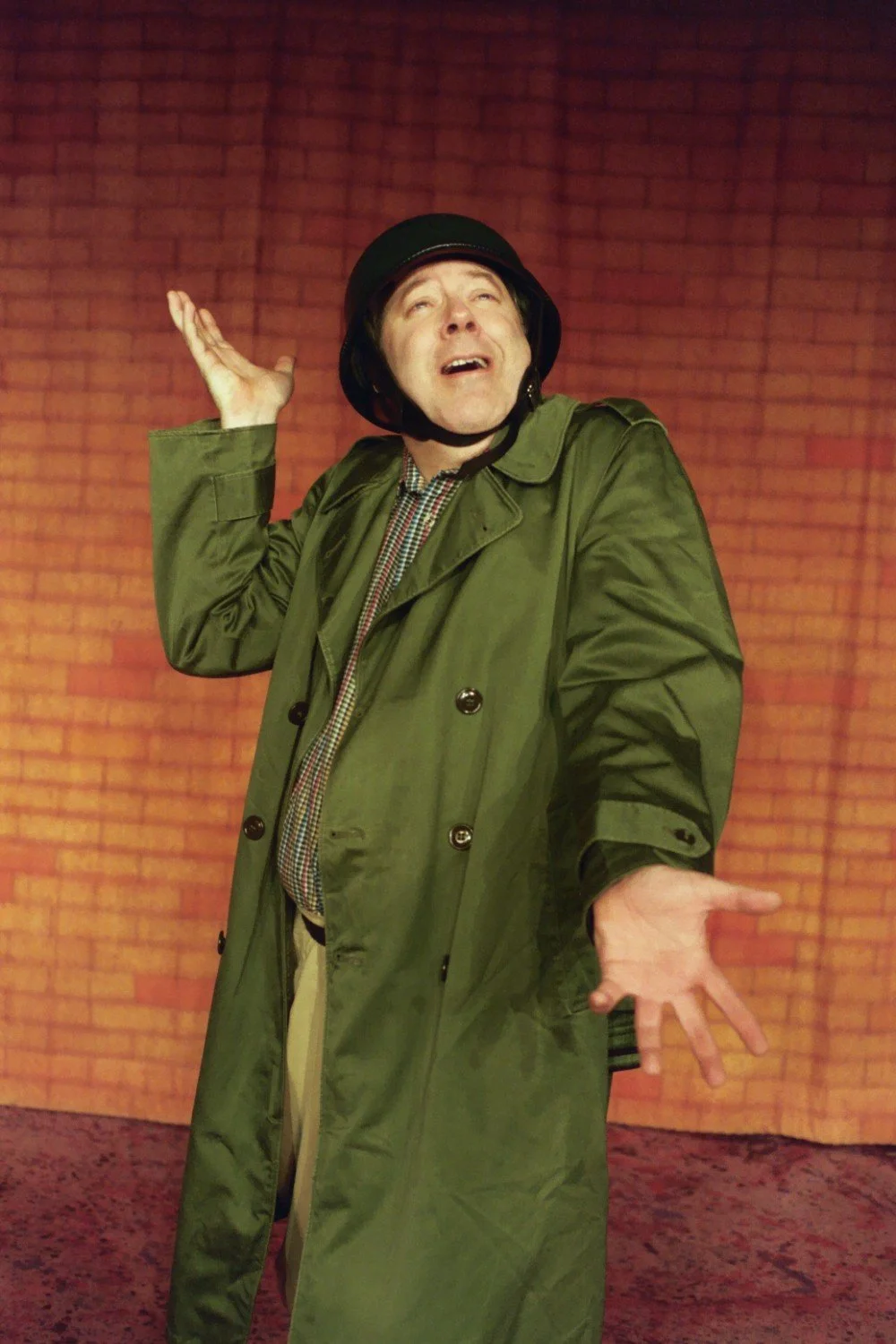 Person in a green trench coat and helmet gesturing with hands against a brick wall background.