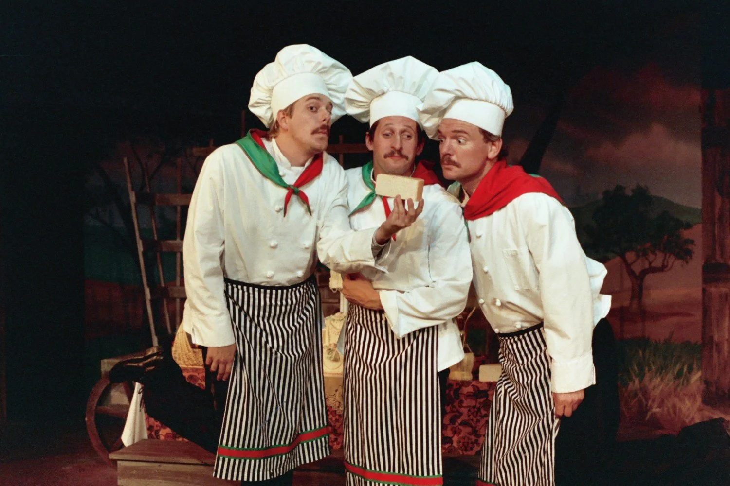 Three chefs in white uniforms and hats hold and examine a cheese block.