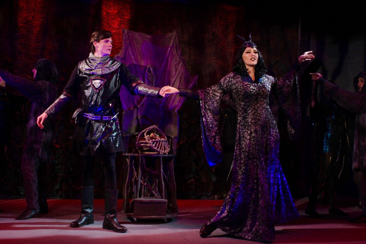 Theatrical performance scene with actors in fantasy costumes, one in dark armor and the other in a long, shimmering gown with a headpiece, set against a dark, atmospheric backdrop.