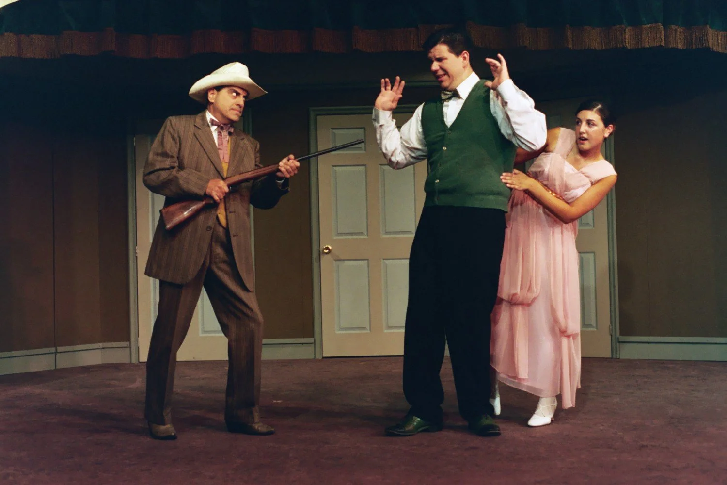 Three actors performing a comedic scene on stage, one wearing a cowboy hat holding a rifle.