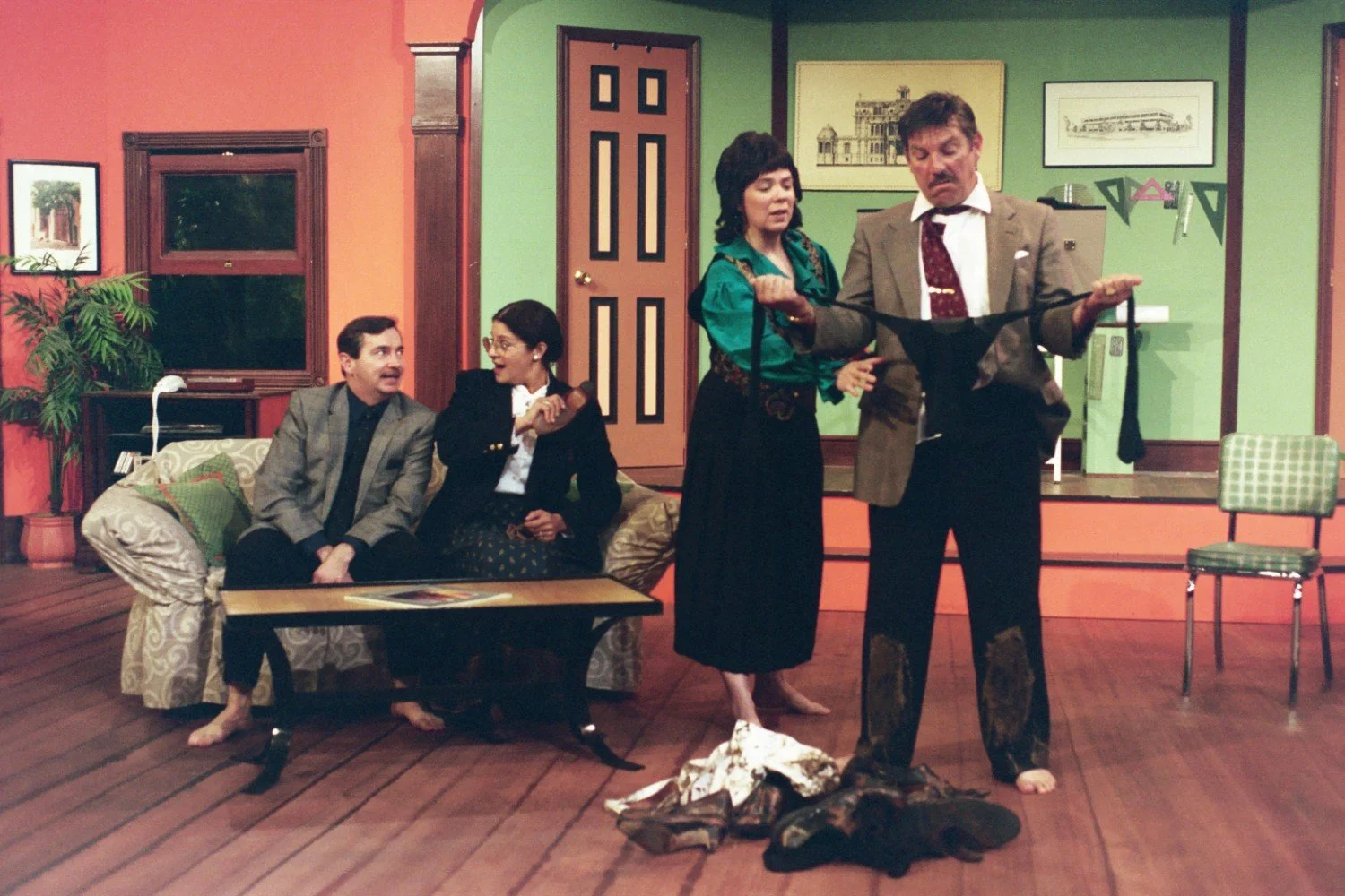 Stage scene with four actors in a colorful living room set, including a sofa, coffee table, and wall art. Two actors are on the sofa conversing; two are standing, holding a piece of clothing.
