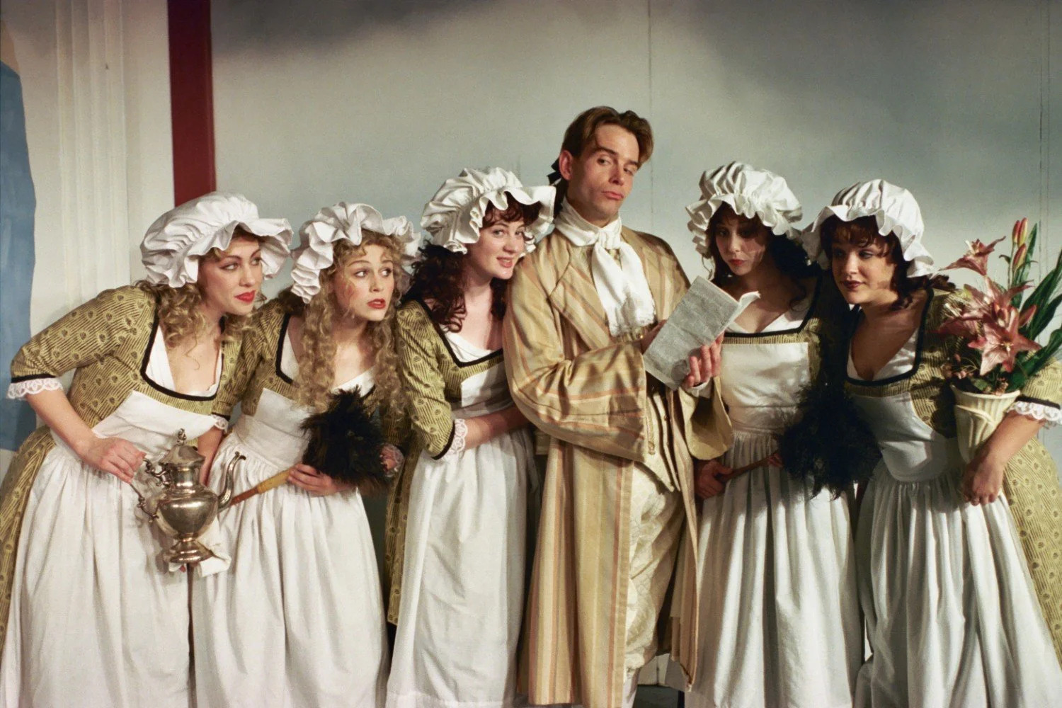 A group of actors in period costumes, featuring a man reading a script and several women wearing maid outfits with bonnets, performing in a theatrical setting.