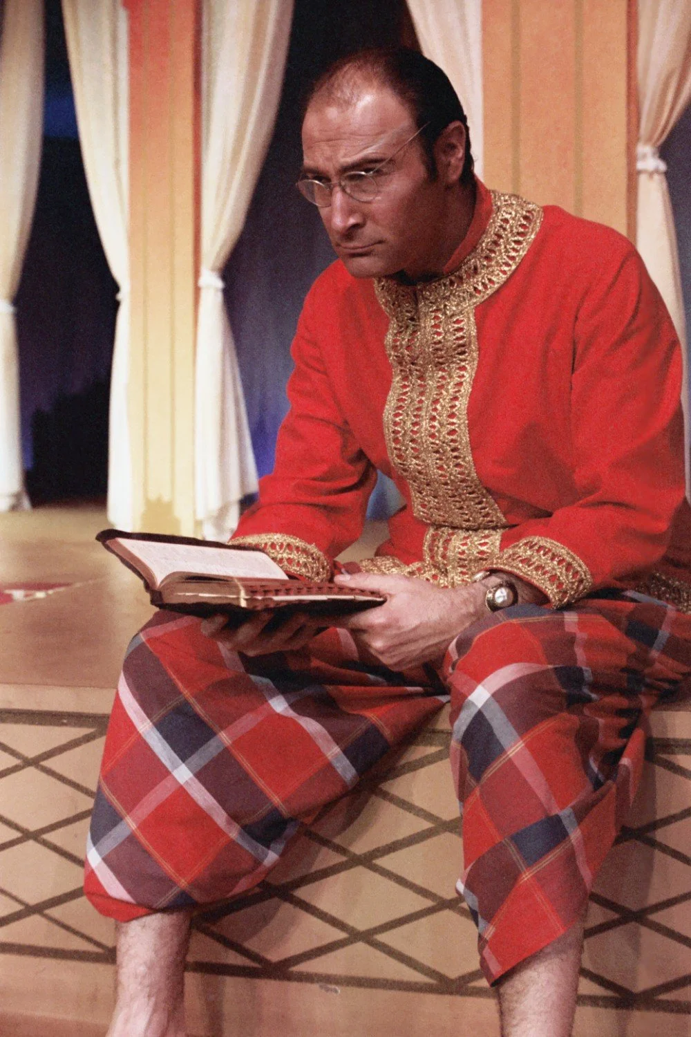 A person wearing a red and gold embroidered outfit, holding a book. The individual is seated and appears to be in a theatrical or performance setting with draped curtains in the background.