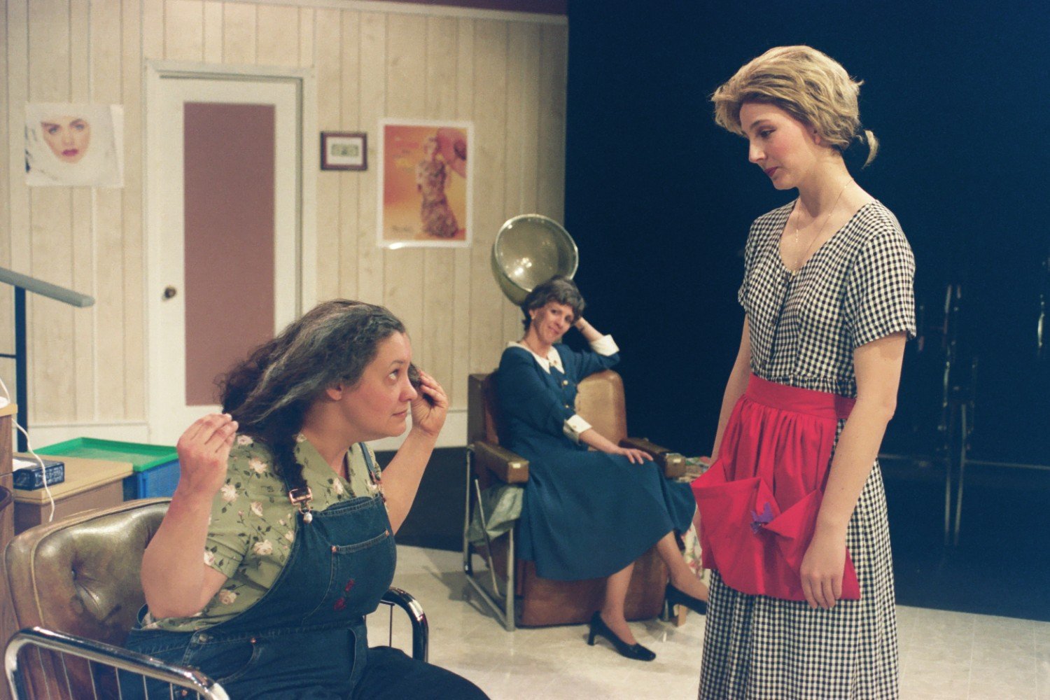 Three women in a vintage-style beauty salon; one seated with long hair, another standing in a gingham dress, and a third sitting under a hairdryer.