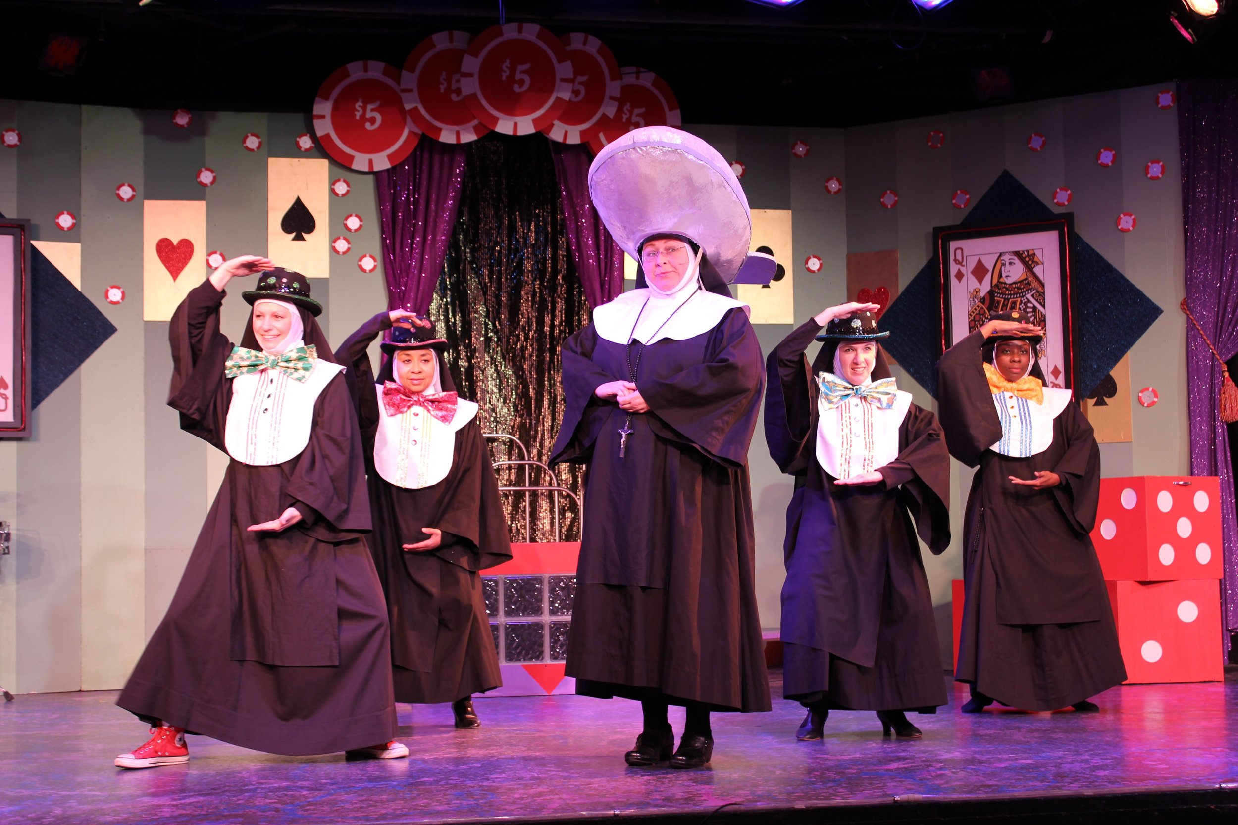 Group of performers dressed as nuns, with exaggerated hats and bow ties, on a stage with a casino-themed background.