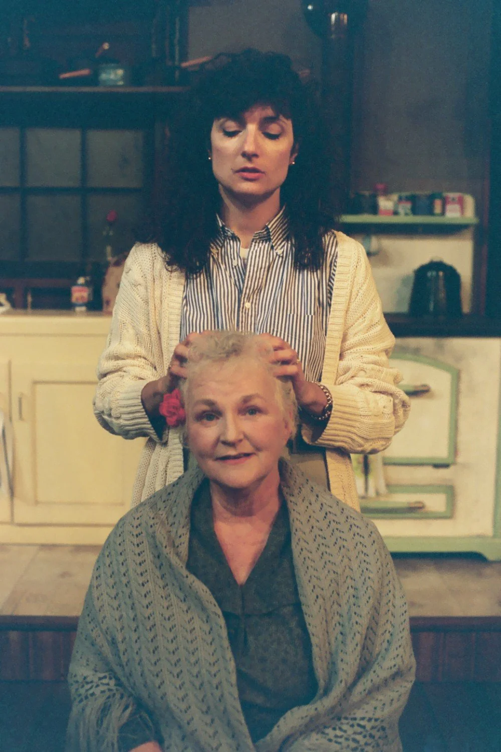 Two women in a vintage kitchen setting, one standing behind the other with hands on her head. The seated woman is smiling and wearing a shawl.
