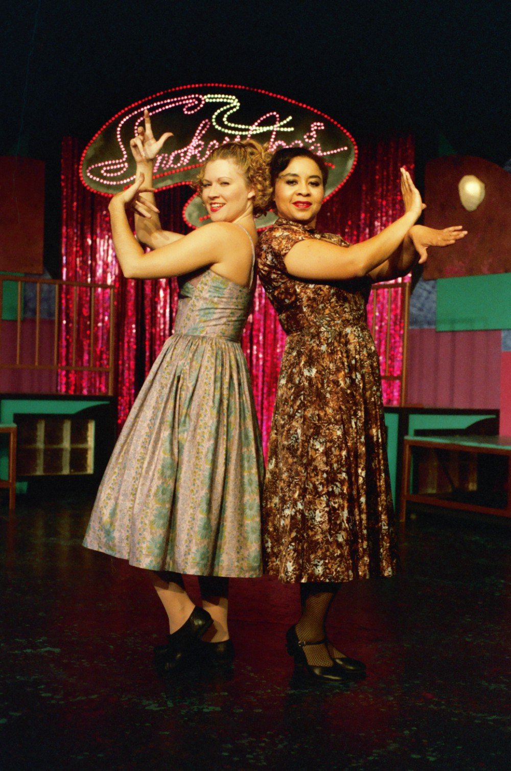 Two women in vintage dresses posing back-to-back, performing a dance move on stage in a retro-themed setting with red curtains and a sign overhead.