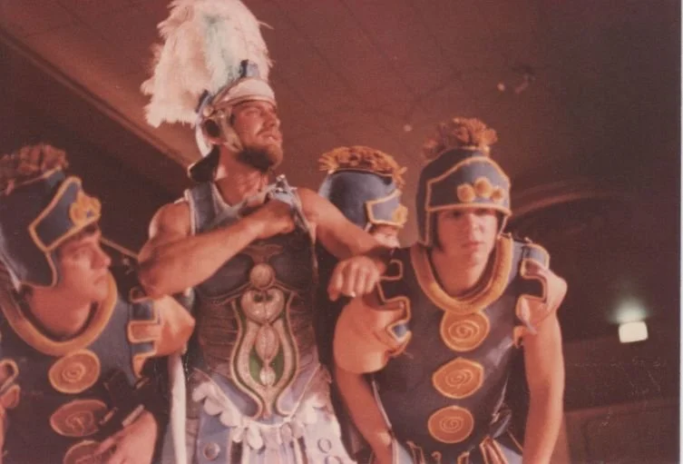 Group of people in stylized Roman or Greek warrior costumes, including helmets with plumes, engaged in a performance.